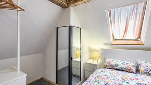 A bedroom with a sloped ceiling, a bed with floral bedding, a bedside table with a lamp, a mirror, and a window with sheer curtains. - Apartment 'Olivias' with Shared Pool, Shared Garden and Wi-Fi (Thürkow)