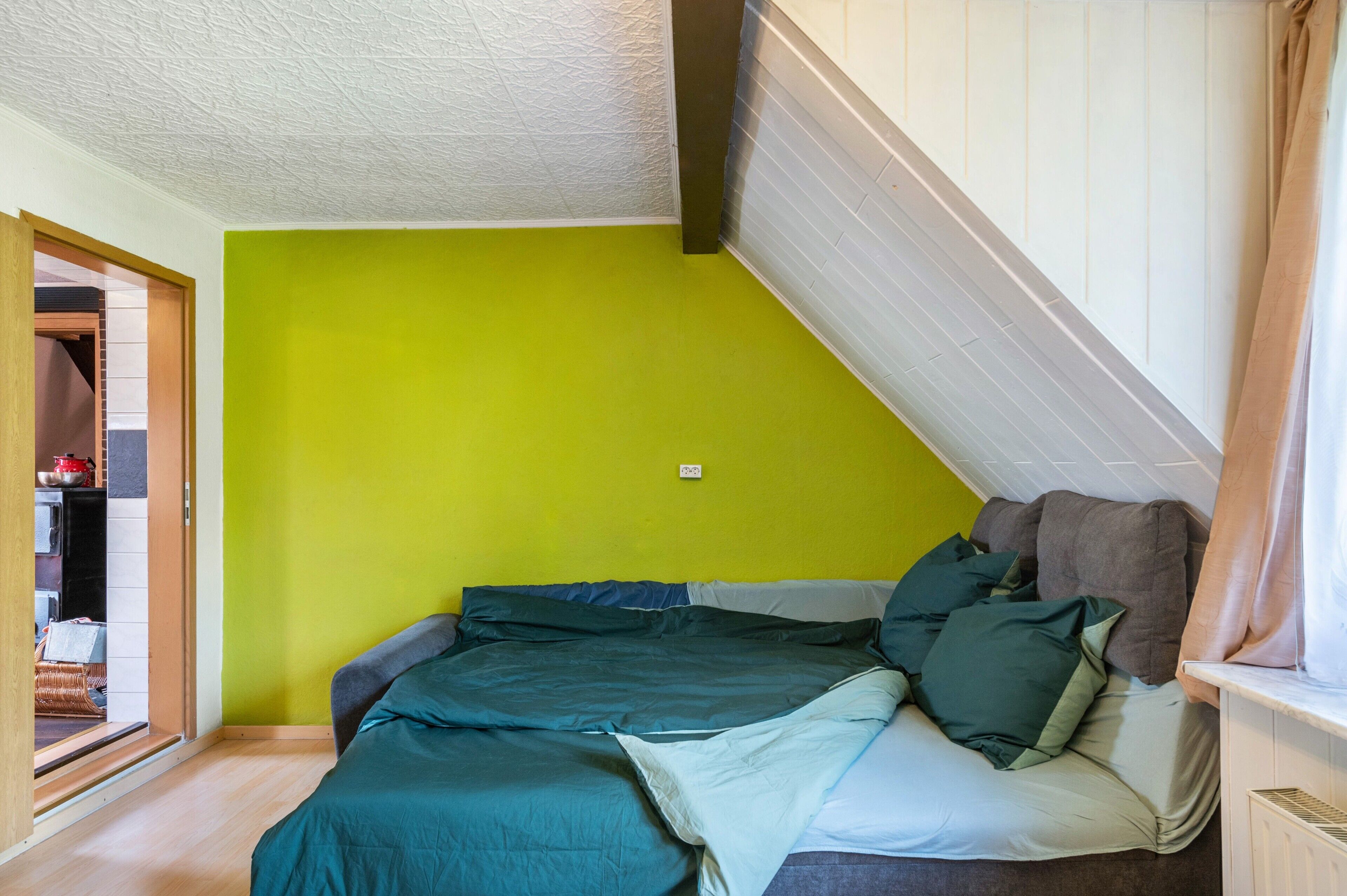 Ein Schlafzimmer mit grüner Wand, einem Bett mit vielen Kissen und einem Fenster mit Vorhängen.