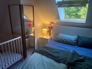 Quarto com berço, mesa de cabeceira com luminária e flores, cama com almofadas e janela com vista para o verde.