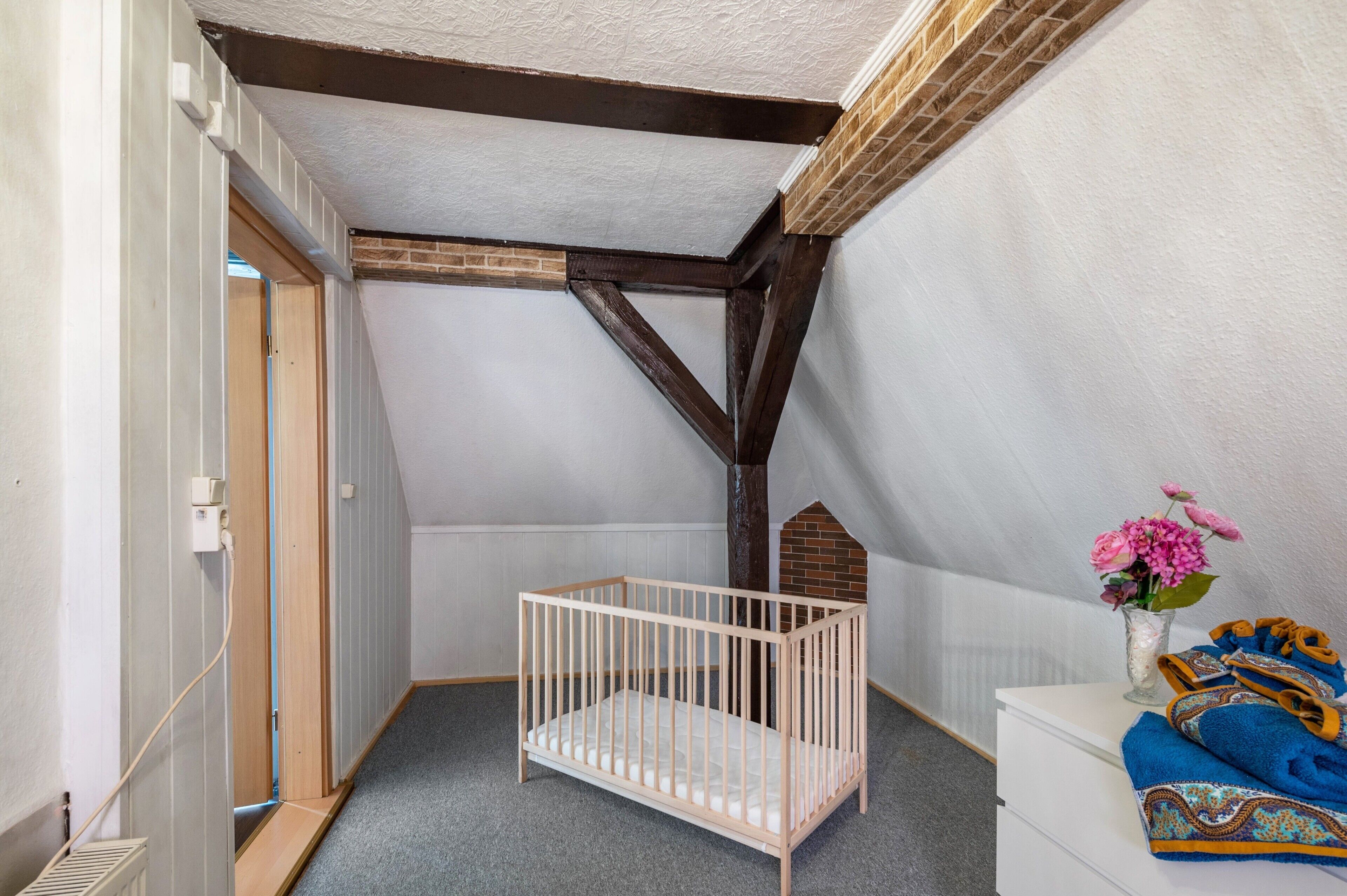 Ein Zimmer mit schrägen Wänden, einem hölzernen Babybett, einer weißen Kommode und einer Vase mit Blumen.