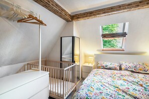 Un dormitorio con una cuna, una cama con ropa de cama floral, un armario y una ventana con vistas a la vegetación.