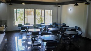 A room with multiple tables and chairs, large windows with blinds, ceiling fans, and a view of greenery outside.