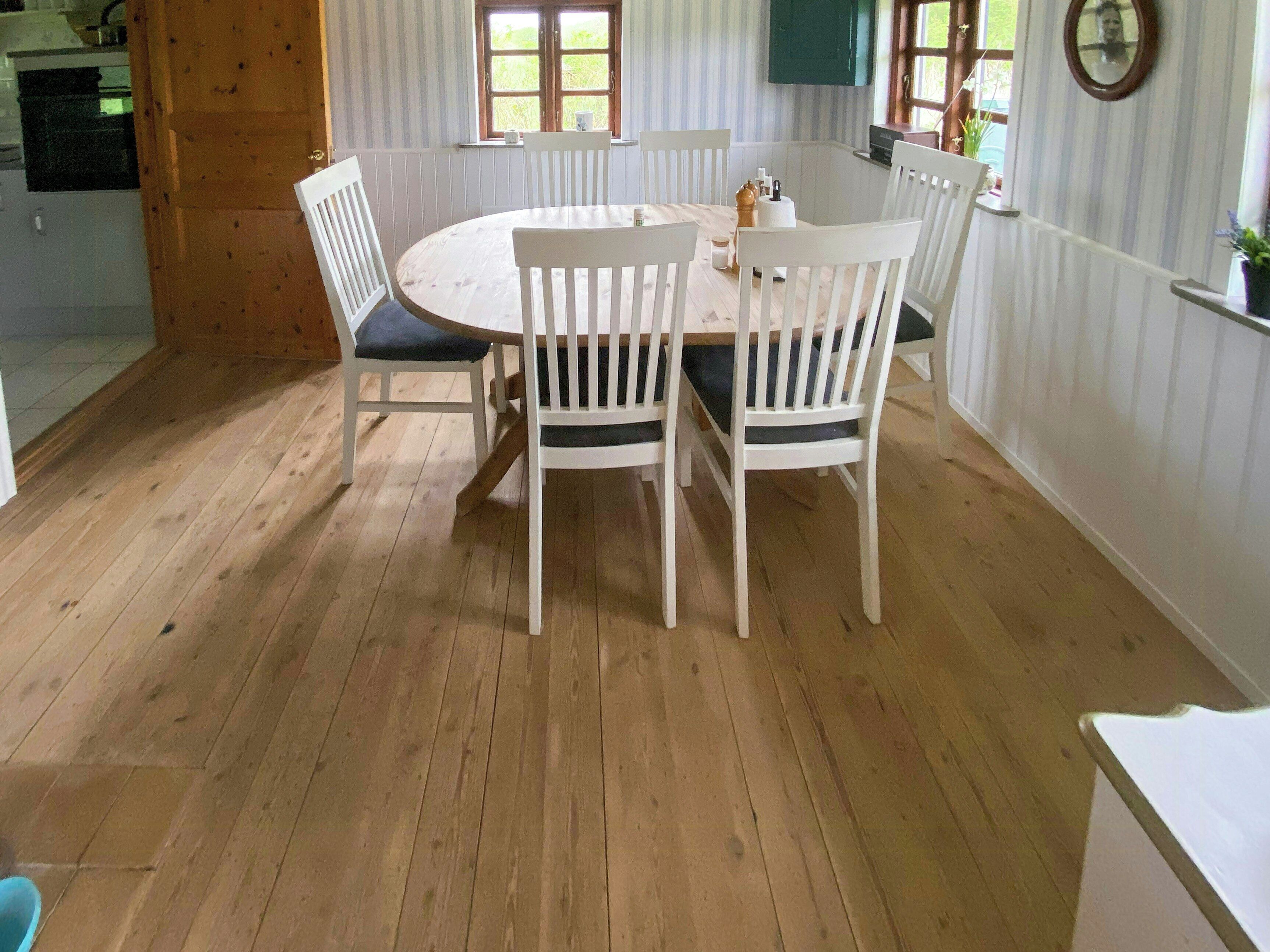 A dining area with a round table and white chairs, wooden flooring, and a window with a view of greenery.