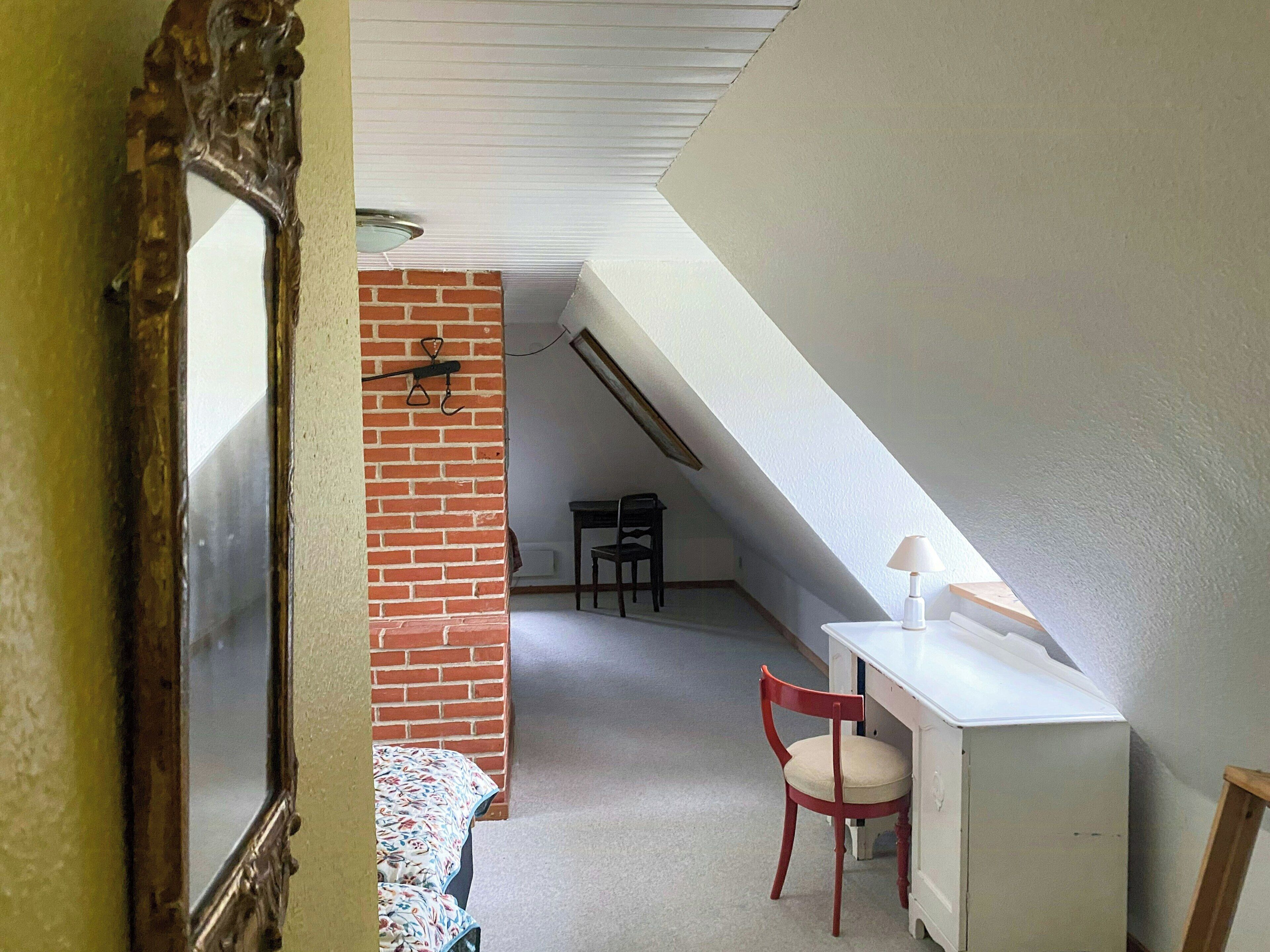 Una habitación con techo inclinado, pared de ladrillo, un escritorio con lámpara, una silla y una cama con colcha estampada.
