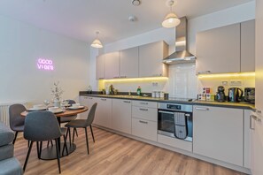 A modern kitchen with white cabinets, a built-in oven, and a dining area with grey chairs. - Stylish 2BR Wembley Stay |Sleeps 8| 5-Min Walk to Stadium (London)