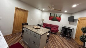 Cocina compacta con comedor, ventilador de techo, televisor montado y sofá rojo.