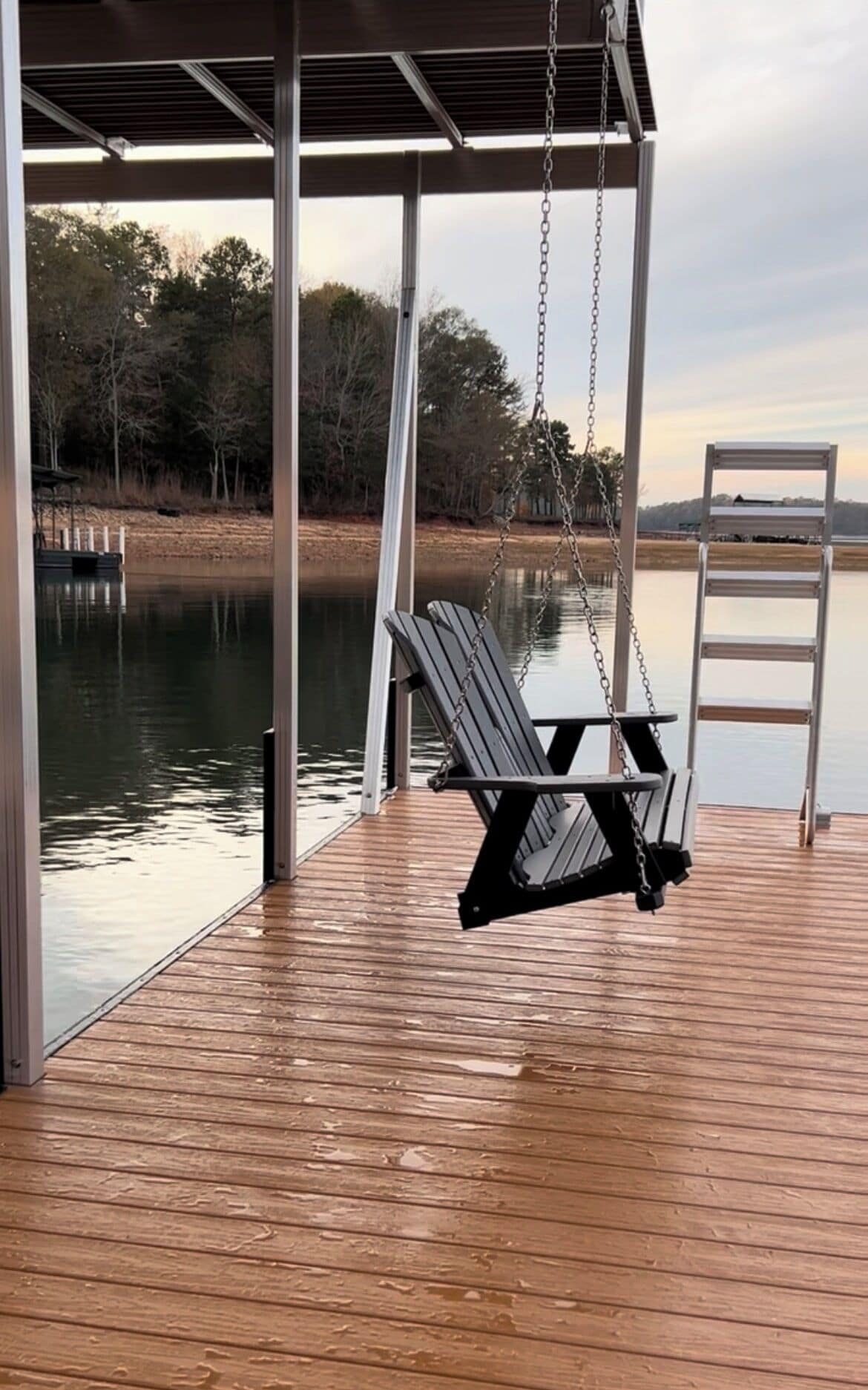 A wooden deck with a swing chair and a ladder, overlooking a calm body of water and trees.