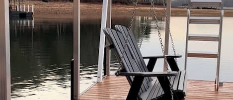 A wooden deck with a swing chair and a ladder, overlooking a calm body of water and trees.