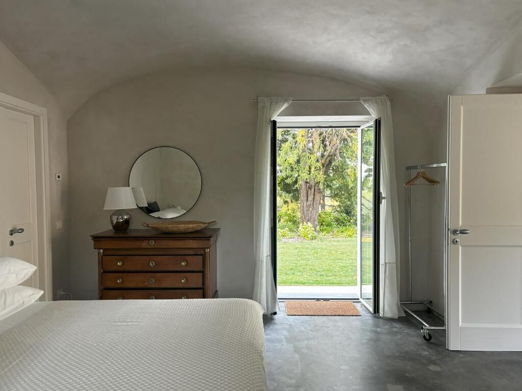 A bedroom with a bed, a dresser with a mirror, a hanging lamp, and a view of a garden through an open door.