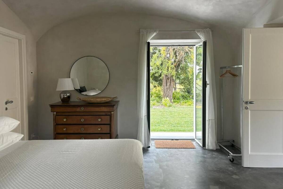 A bedroom with a bed, a dresser with a mirror, a hanging lamp, and a view of a garden through an open door.