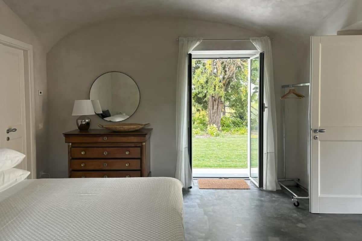 Un dormitorio con cama, cómoda con espejo, lámpara colgante y vistas a un jardín a través de una puerta abierta.