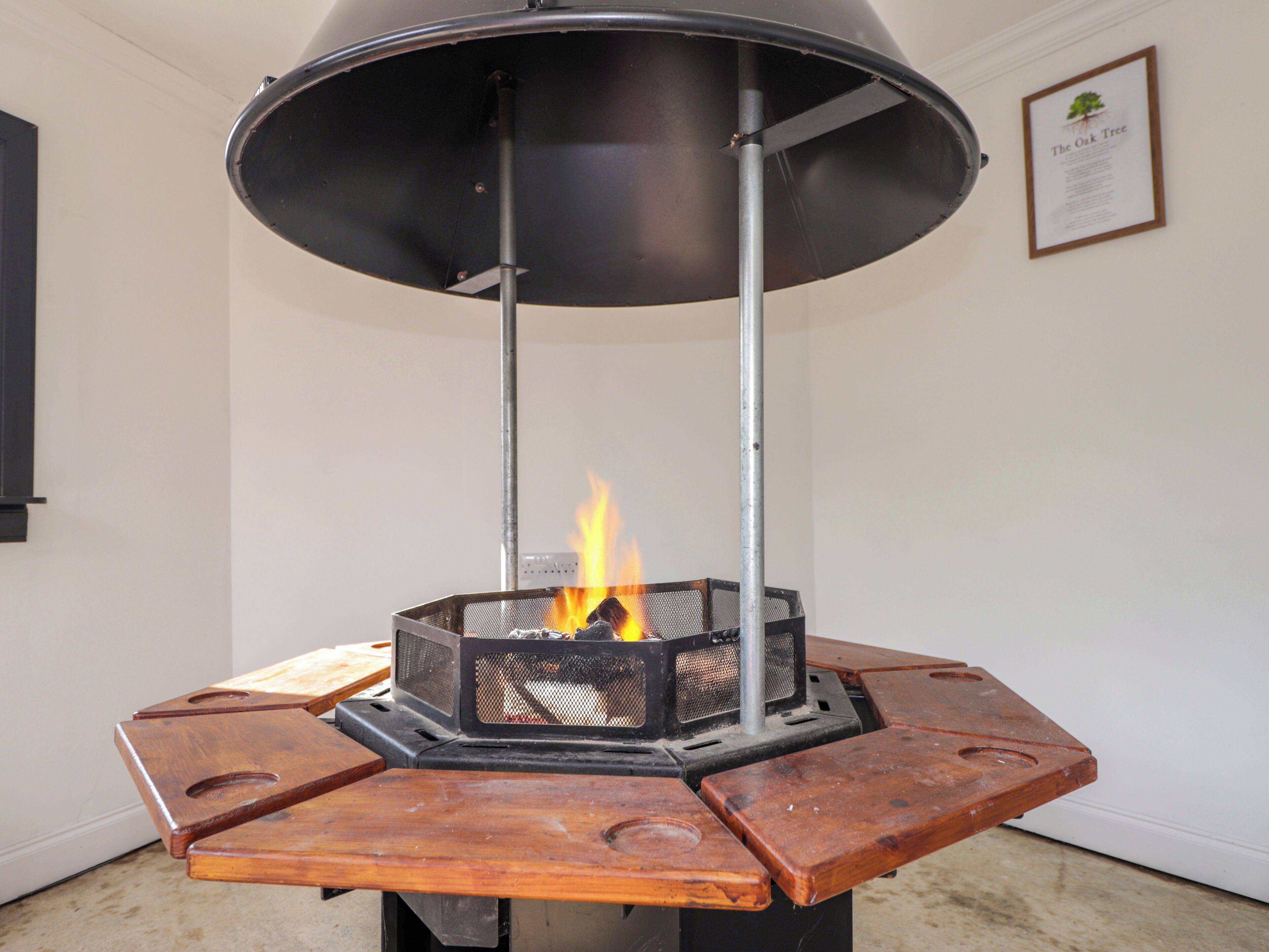 Un fogón de leña con estructura metálica y parte superior negra, colocado sobre una plataforma de madera con fuego ardiendo en su interior.