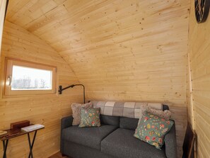 A cozy cabin interior with a wooden ceiling, a sofa with patterned cushions, a small table with a book, and a window with a view.