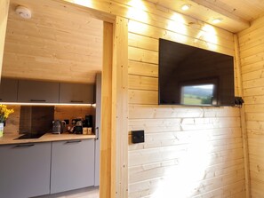 A wooden interior with a kitchen area, including cabinets, a countertop, and a wall-mounted television. - Little Oaks (Dingwall)