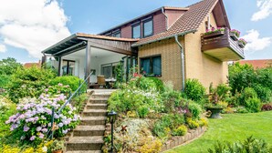 Et toetagers hus med en overdækket veranda, en murstensfacade og en have med forskellige planter og blomster.