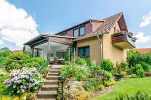 A two-story house with a covered porch, a brick exterior, and a garden with various plants and flowers. - Koch Holiday Apartment up to 100m² with Balcony in Bad Grund (Bad Grund (Harz))