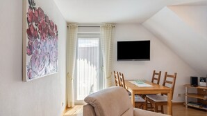 En stue med en beige sofa, et spisebord med stole, et fladskærms-tv monteret på væggen og et vindue med gardiner.