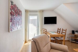A living room with a beige sofa, a dining table with chairs, a flat-screen TV mounted on the wall, and a window with curtains. - Koch Holiday Apartment up to 100m² with Balcony in Bad Grund (Bad Grund (Harz))
