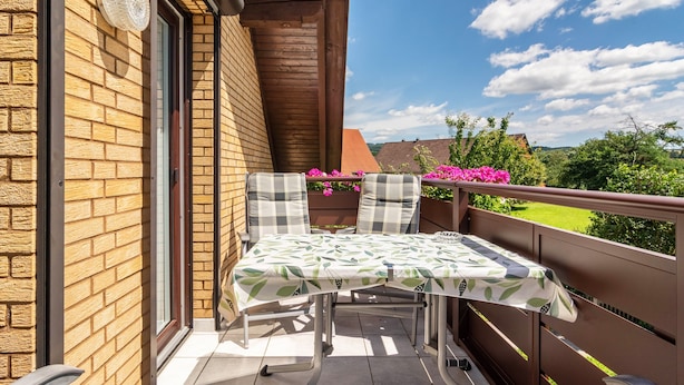 En balkon med et bord og stole, en murstensvæg, gelænder i træ og udsigt til grønt og huse.
