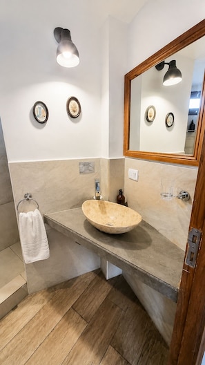Un baño con piso de madera, pared de azulejos color beige, un lavamanos redondo, toallero y espejo con marco de madera.
