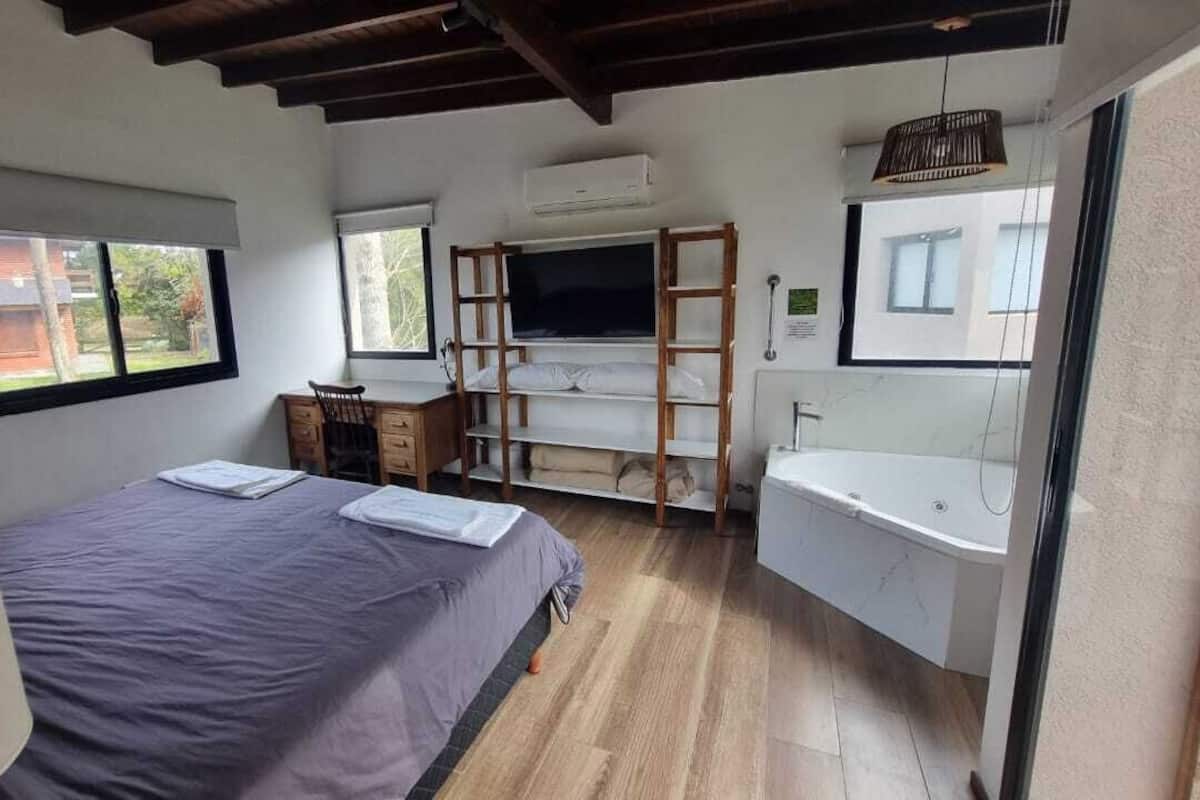 Quarto com cama, escrivaninha, televisão, prateleira, janela e banheiro com banheira.