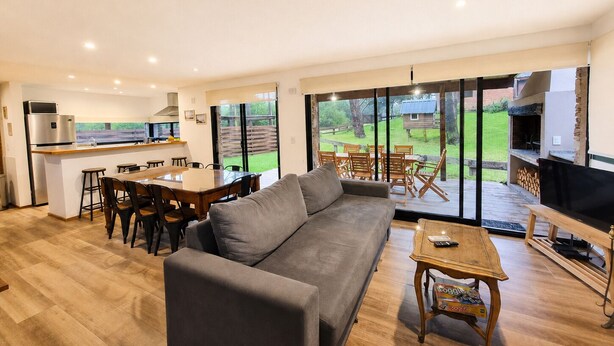 A modern living room with a sofa, dining table, and chairs, a kitchen area, and a large sliding glass door leading to a backyard with outdoor furniture. - Complejo Playa Bonita Mar Azul 100m playa (Mar Azul)