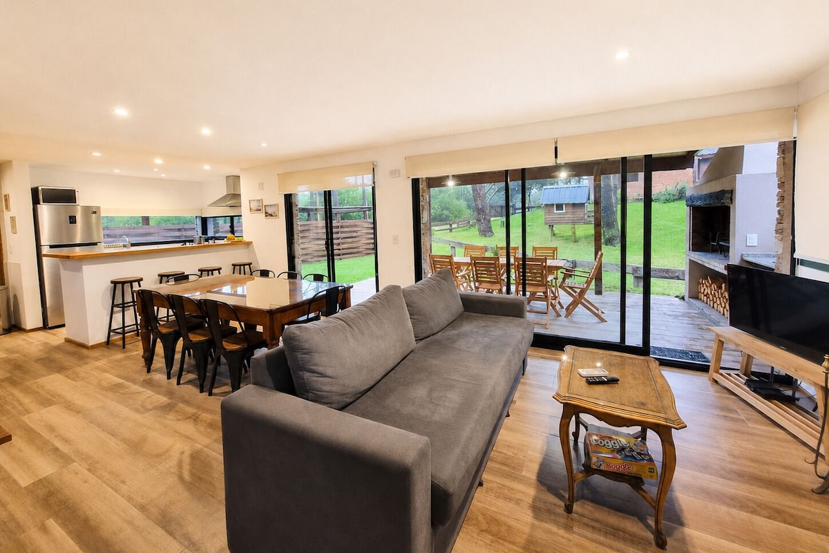 Una sala de estar moderna con sofá, mesa de comedor y sillas, zona de cocina y un gran ventanal corredero que da a un patio trasero con mobiliario exterior.