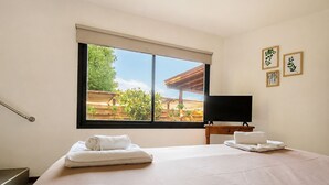 Habitación de hotel con una cama grande, un televisor sobre una mesita de madera y una ventana con vistas a una terraza de madera y vegetación.
