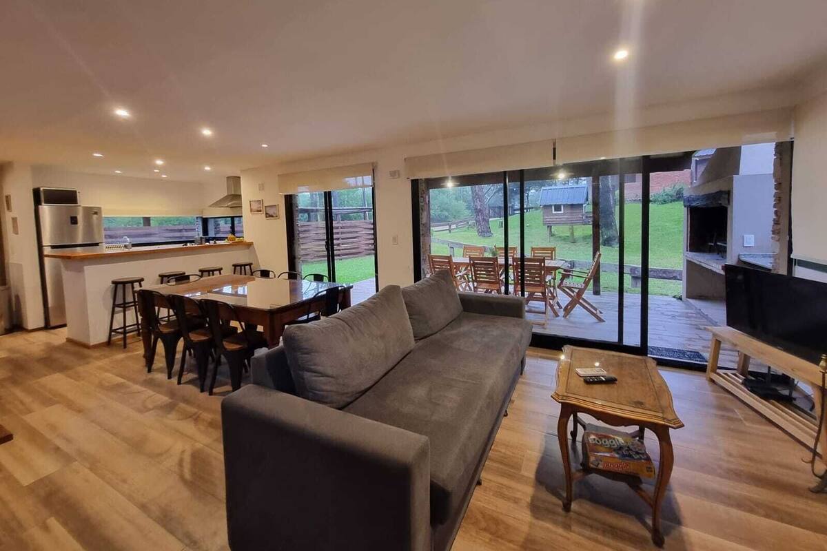 Amplia sala de estar con un sofá gris, una mesa de comedor de madera y sillas, un área de cocina con refrigerador y un patio con parrilla y mobiliario exterior.