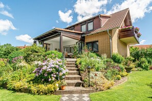 Ein zweistöckiges Haus mit überdachter Terrasse, Backsteinfassade und einem Garten mit verschiedenen Pflanzen und Blumen.