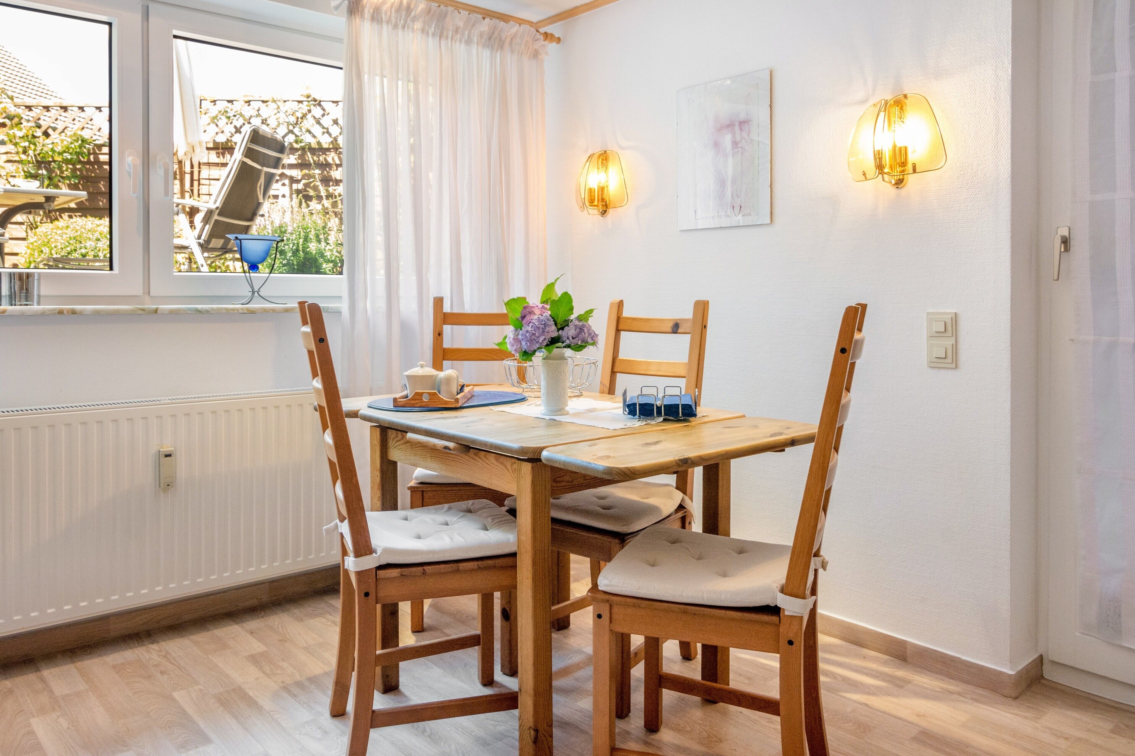 Un coin repas avec une table et des chaises en bois, un vase de fleurs et des lampes fixées au mur.