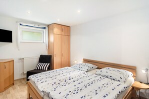 Ein Schlafzimmer mit einem Bett, einem Sessel, einem Kleiderschrank, einer Heizung, einem Fenster und einem an der Wand befestigten Fernseher.