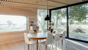 Een moderne eetruimte met een houten tafel, witte stoelen, een zwarte hanglamp en grote ramen met uitzicht op bomen.