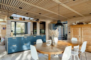 Et moderne køkken med træloft, en central ø med blå skabe, og et spiseområde med et træbord og hvide stole.
