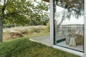 Et moderne udendørs opholdsområde med glasfacade, træterrasse og udsigt til bro og træer.