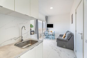 Une cuisine moderne avec un plan de travail et un évier en marbre, un coin repas avec des chaises bleues et un canapé gris sur lequel est fixé un téléviseur au mur.