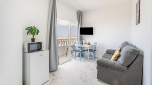 A modern living room with a sofa, a TV, a dining table with chairs, a small refrigerator, and a balcony with curtains. - Vacation Apartment 'Balcón Panorámico, Mar Y Puesta Sol' with Sea View, Balcony and Wi-Fi (Guargacho)