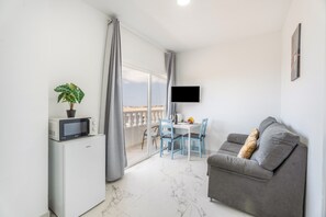 Un soggiorno moderno con divano, televisore, tavolo da pranzo con sedie, un piccolo frigorifero e un balcone con tende.