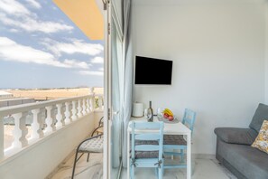 Un balcon avec une table blanche, des chaises bleues, un canapé gris et un téléviseur fixé au mur.