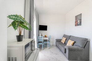 Ein modernes Wohnzimmer mit grauem Sofa, einem Esstisch mit Stühlen, einer Topfpflanze und einem an der Wand montierten Flachbildfernseher.