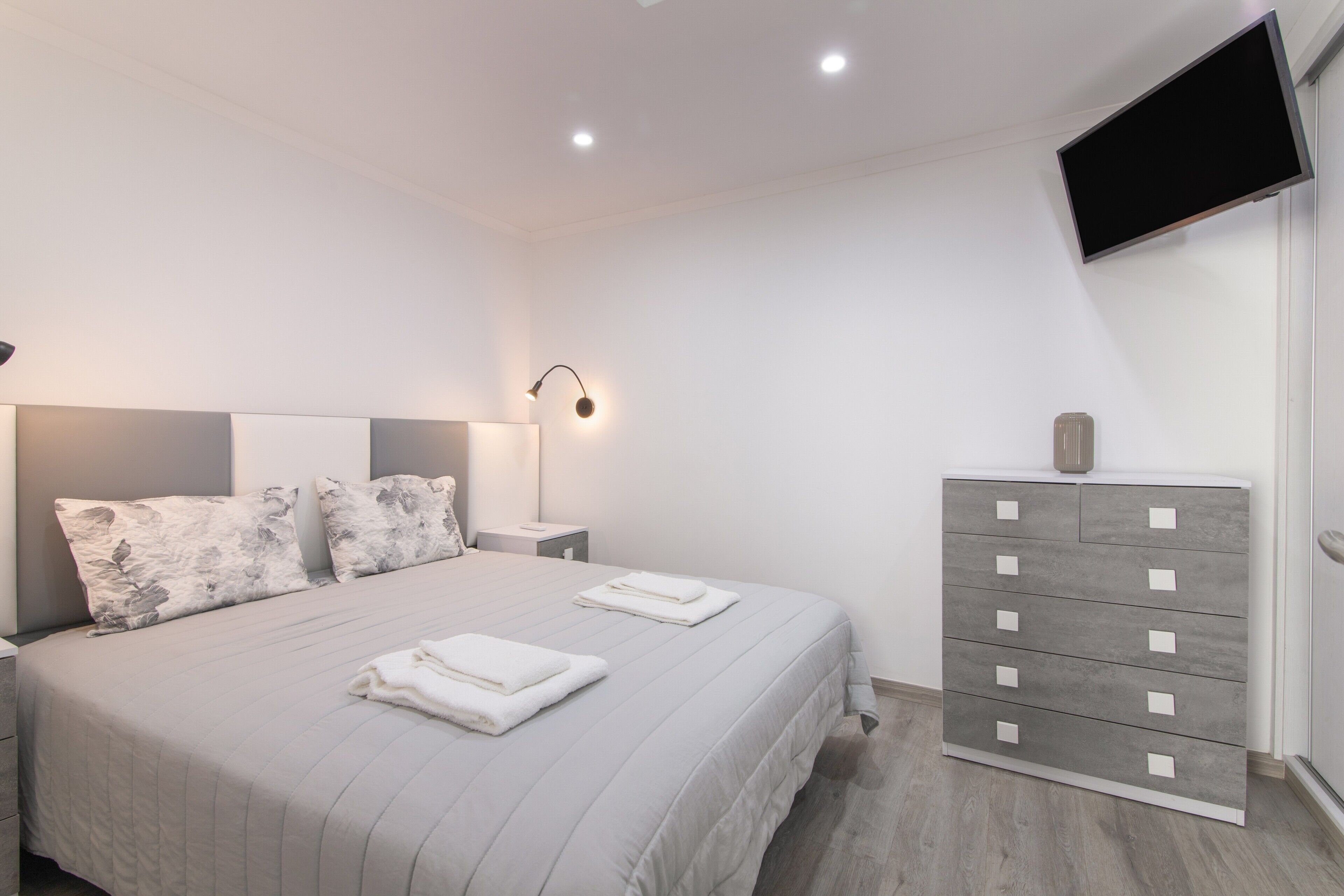 A modern bedroom with a bed, nightstand, dresser, and a flat-screen TV mounted on the wall.