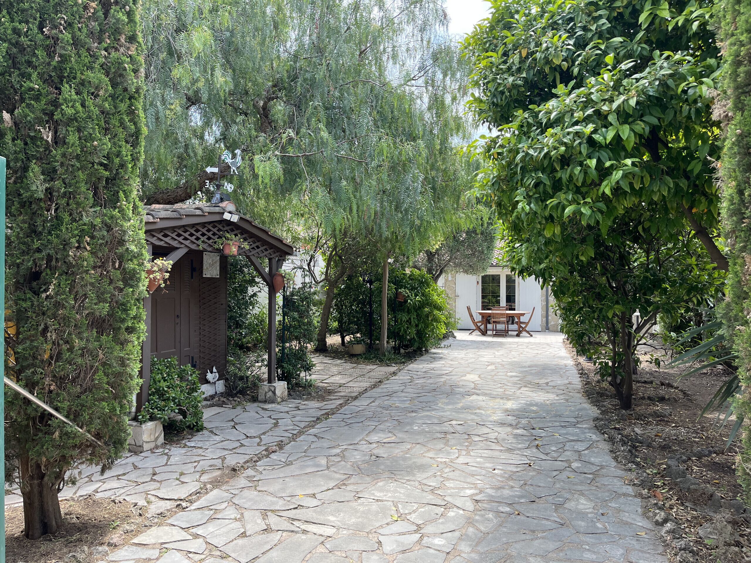 A stone pathway leading to a wooden pergola with a table and chairs, surrounded by lush greenery and a white building in the background.