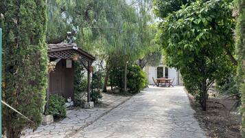 A stone pathway leading to a wooden pergola with a table and chairs, surrounded by lush greenery and a white building in the background.