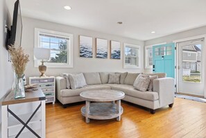 A living room with a sectional sofa, a round coffee table, a TV mounted on the wall, and a door leading outside. - Luxury Coastal Retreat w/ Private Beach Access + Water Views, Clinton, CT (Clinton)