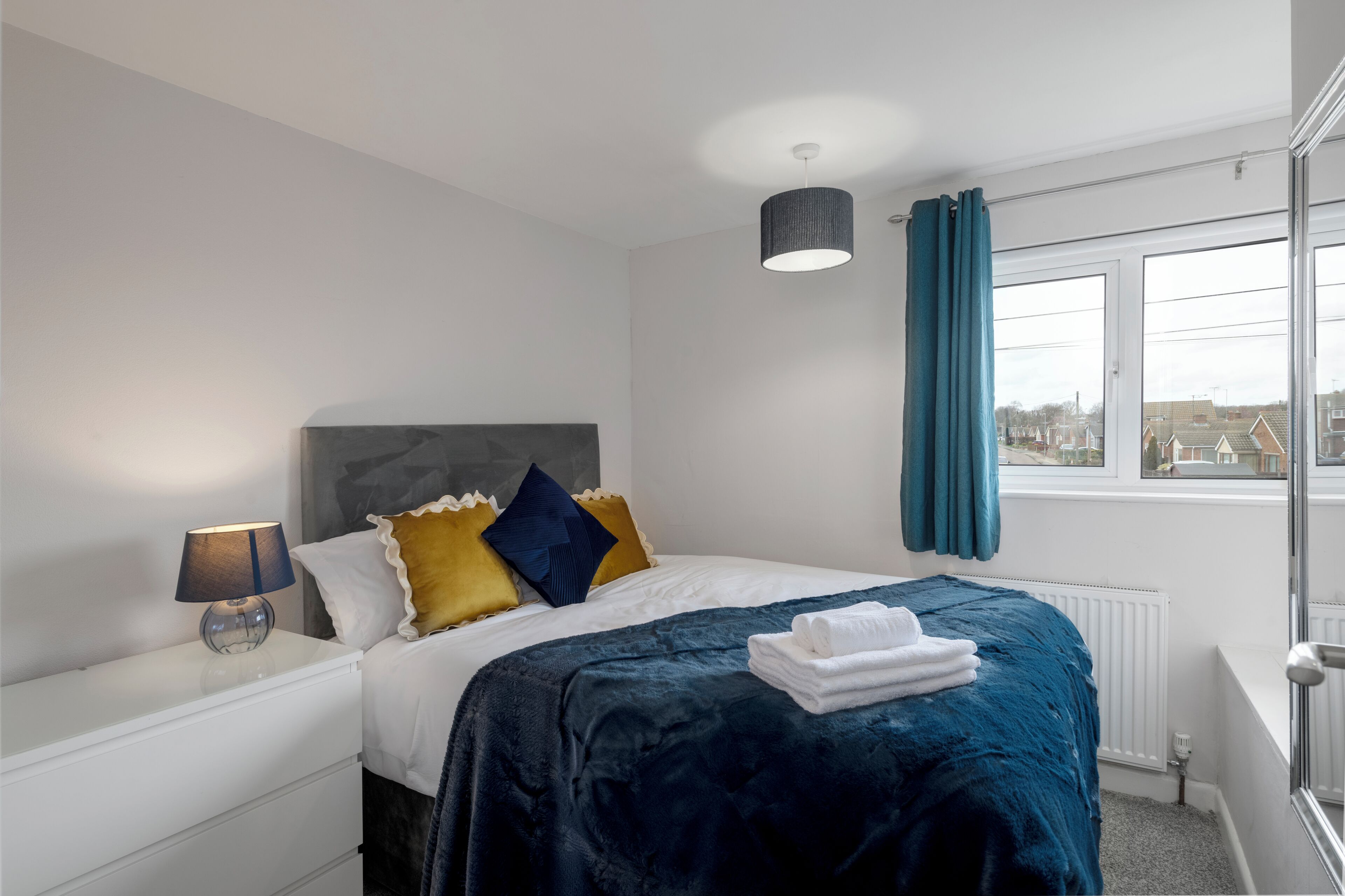 A bedroom with a bed, a nightstand, a lamp, a window with curtains, and a view of buildings outside.