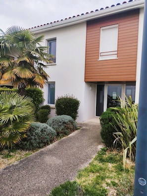 Ein modernes, zweistöckiges Haus mit einem Schotterweg, einem kleinen Garten mit verschiedenen Sträuchern und einer Palme.