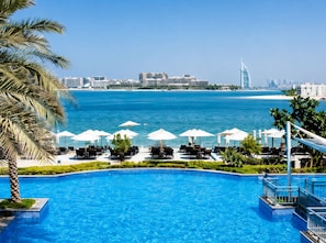 Une piscine avec des chaises longues et des parasols, un palmier, et une vue sur la mer et le paysage urbain.