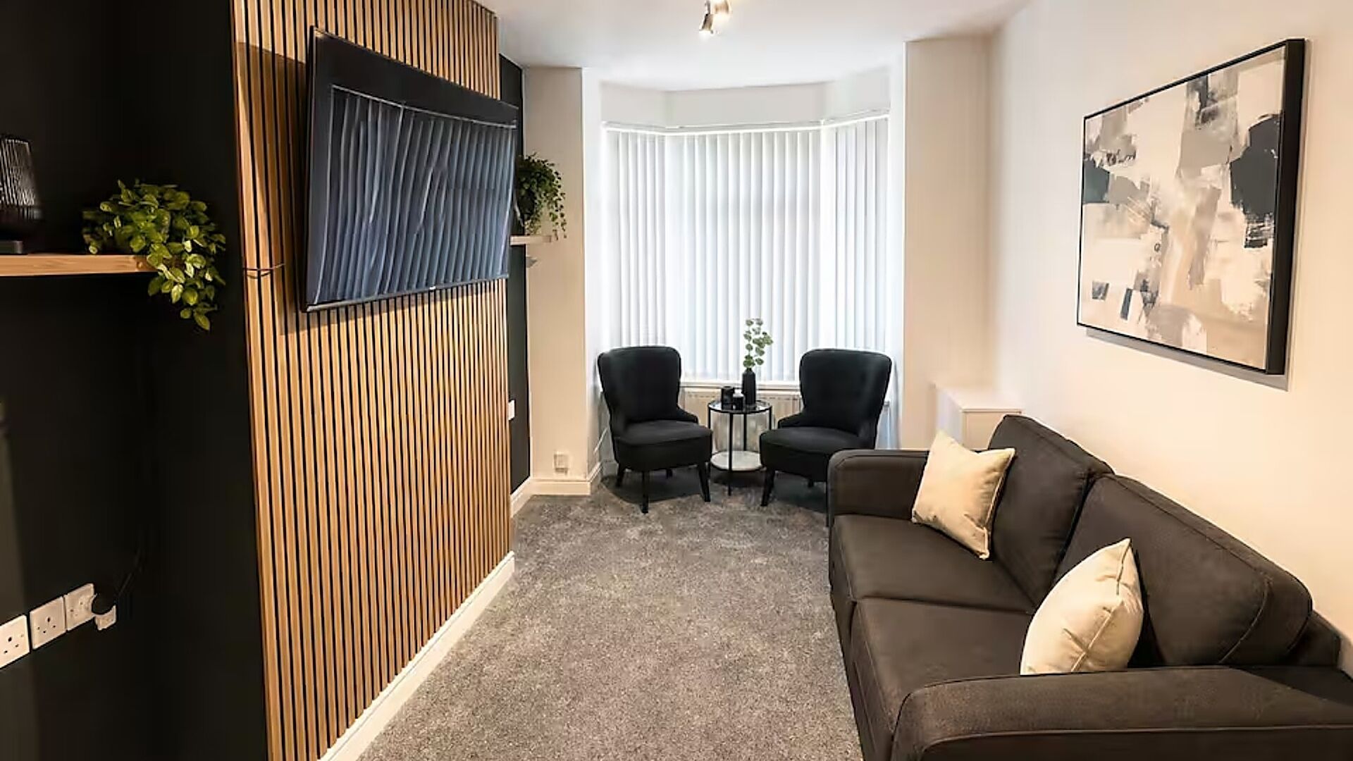 Ein modernes Wohnzimmer mit einem dunkelbraunen Sofa, zwei schwarzen Sessel, einem kleinen runden Tisch und einem großen abstrakten Gemälde an der Wand.