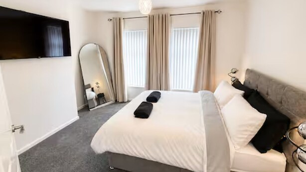 A hotel room with a bed, a TV mounted on the wall, a bedside table with a lamp, and a large mirror.