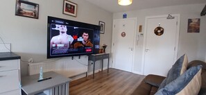 A living room with a flat-screen TV mounted on the wall, a white radiator, a small table with a lamp, a brown sofa, and a door with a wreath. - Cozy 1BDR APT with Private Balcony Near City (Cork)
